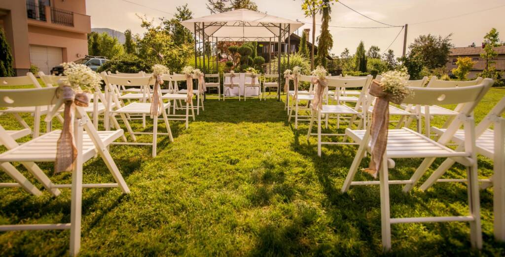 A simple outdoor wedding ceremony setup in a lush green garden with floral decor, aisle seating, and natural surroundings